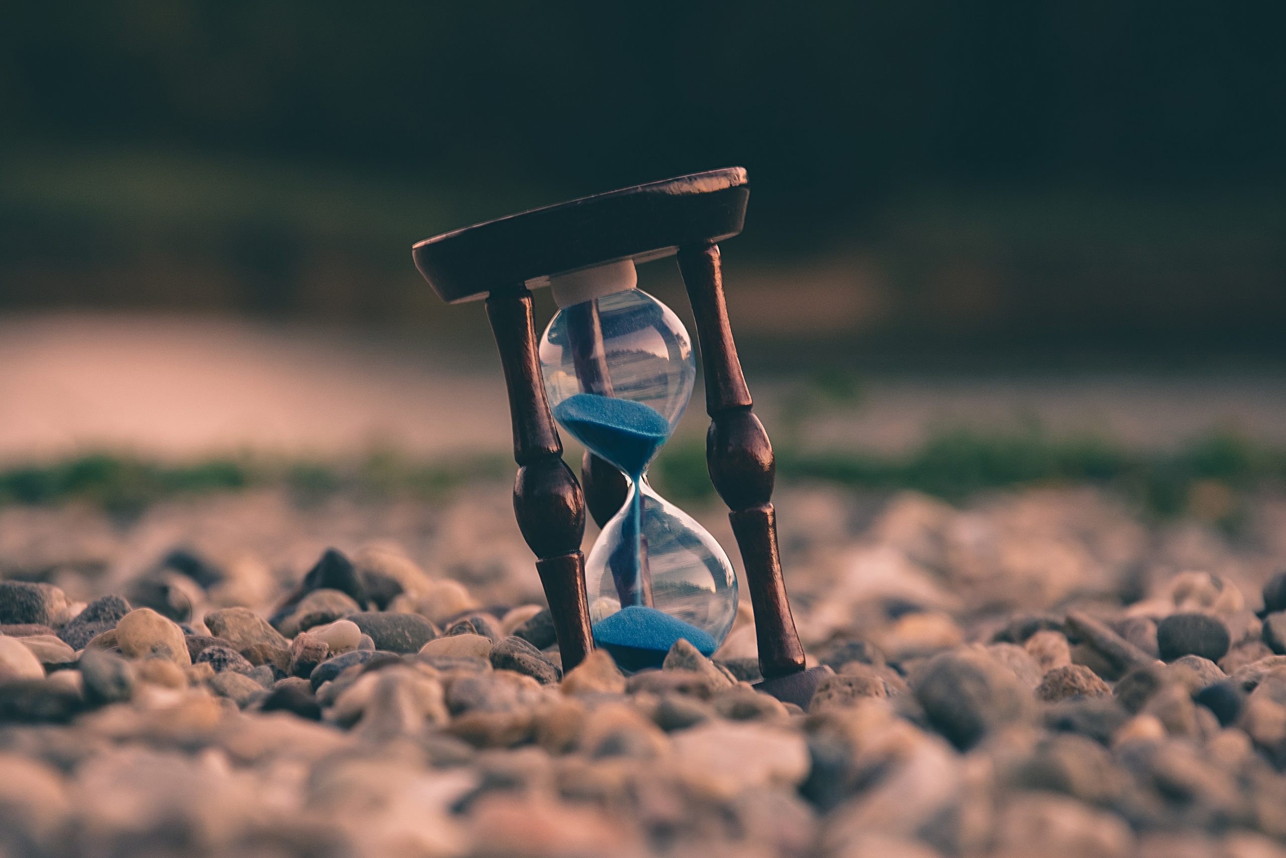an hourglass filled with blue sand sits at a tilt on a bank of pebbles