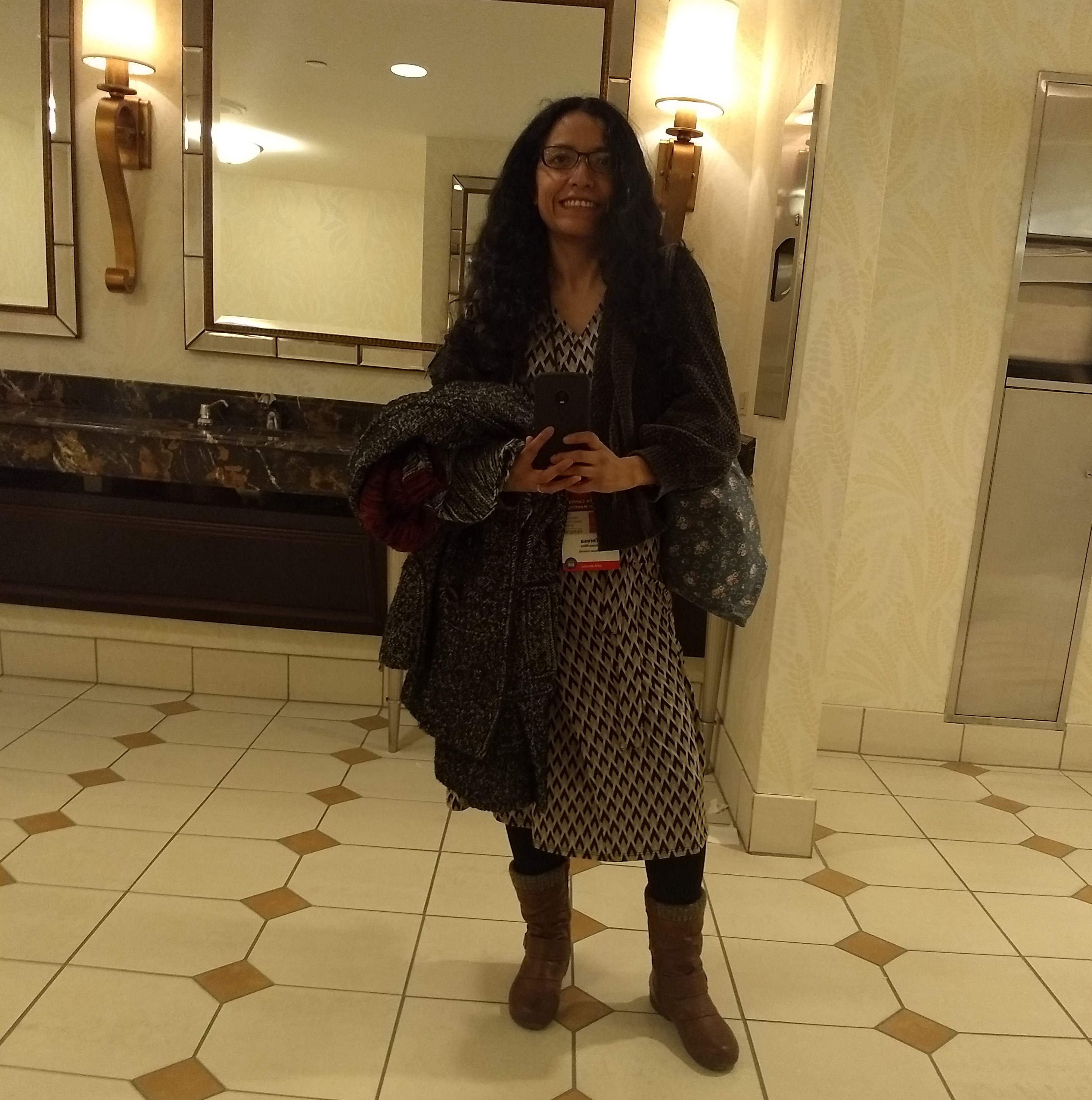 Image of Teresa Bruno Nino reflected in a restroom mirror in the hotel conference at the Central APA 2019 in Chicago. Teresa Bruno Nino is holding her winter coat, taking the selfie and carrying a bag. She has long dark hair. She’s wearing glasses, a dress and boots.  