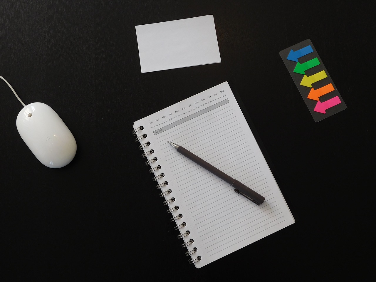 Overhead shot of black desktop with mouse, blank agenda and pen, flashcards, and sticky notes on it