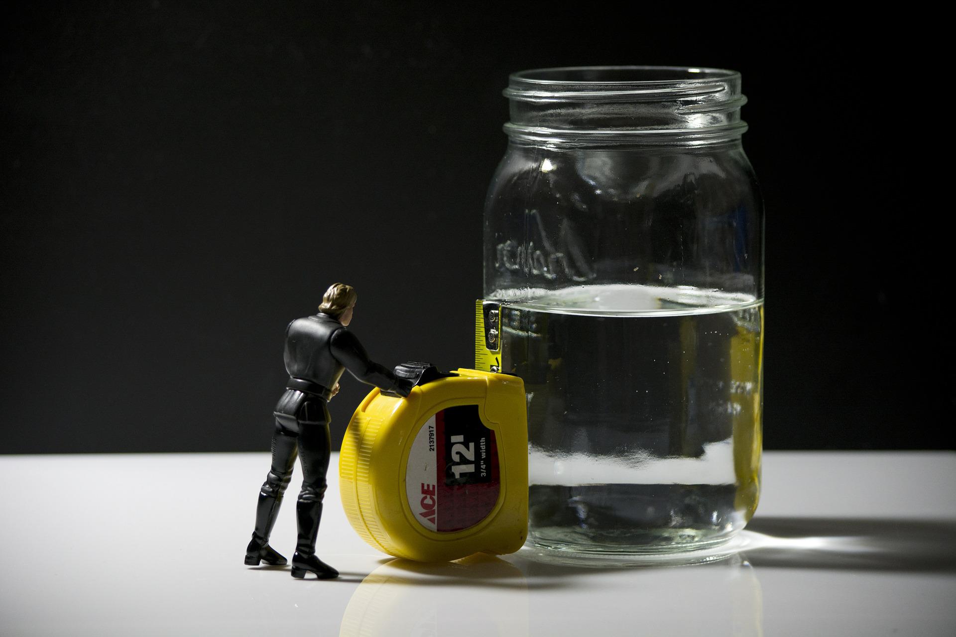 Toy man measuring water level in glass with tape measure - is it half full or half empty?