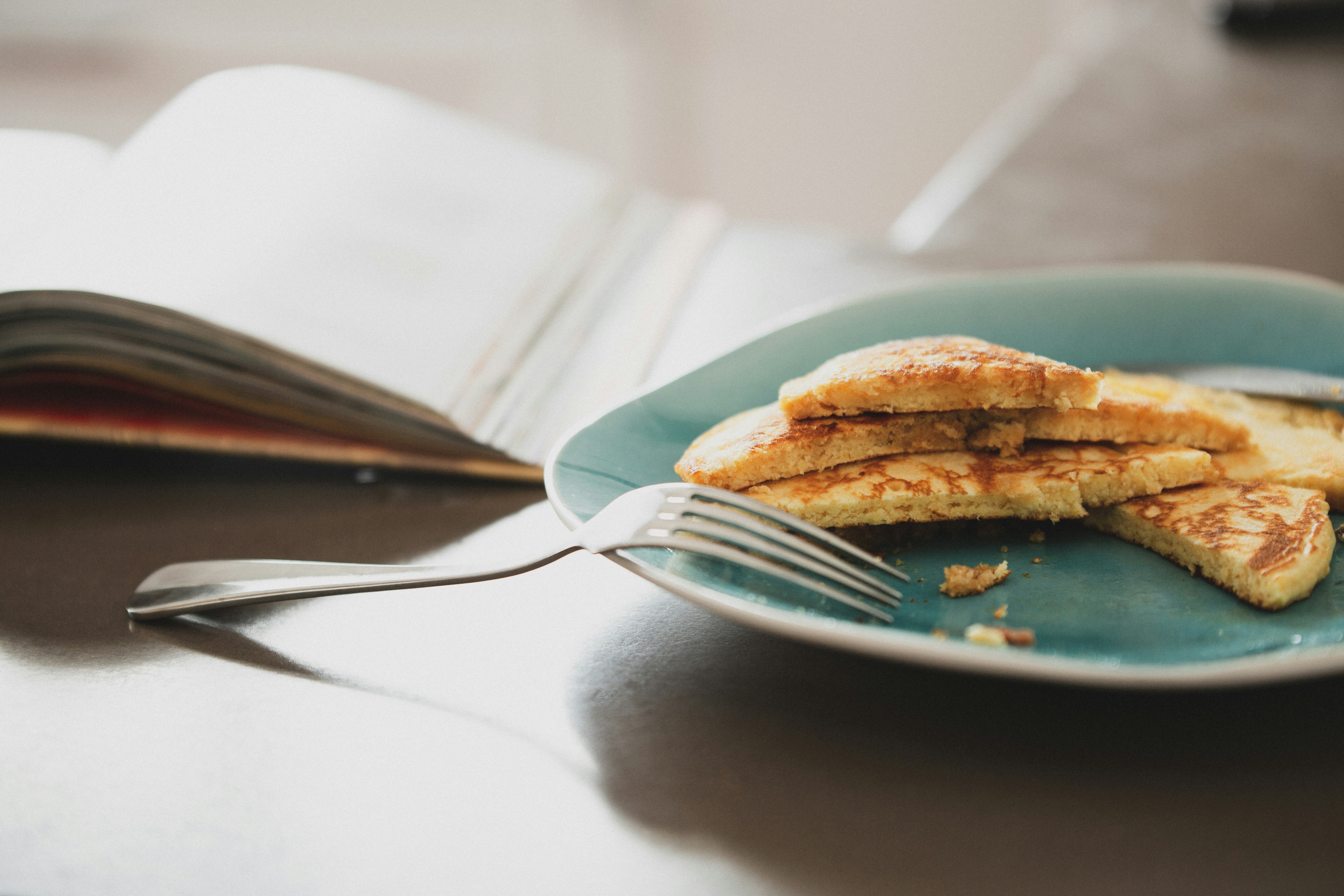 On a table sits a blue plate with a stack of pancake half eaten, an overturned fork, and a book open but out of focus.