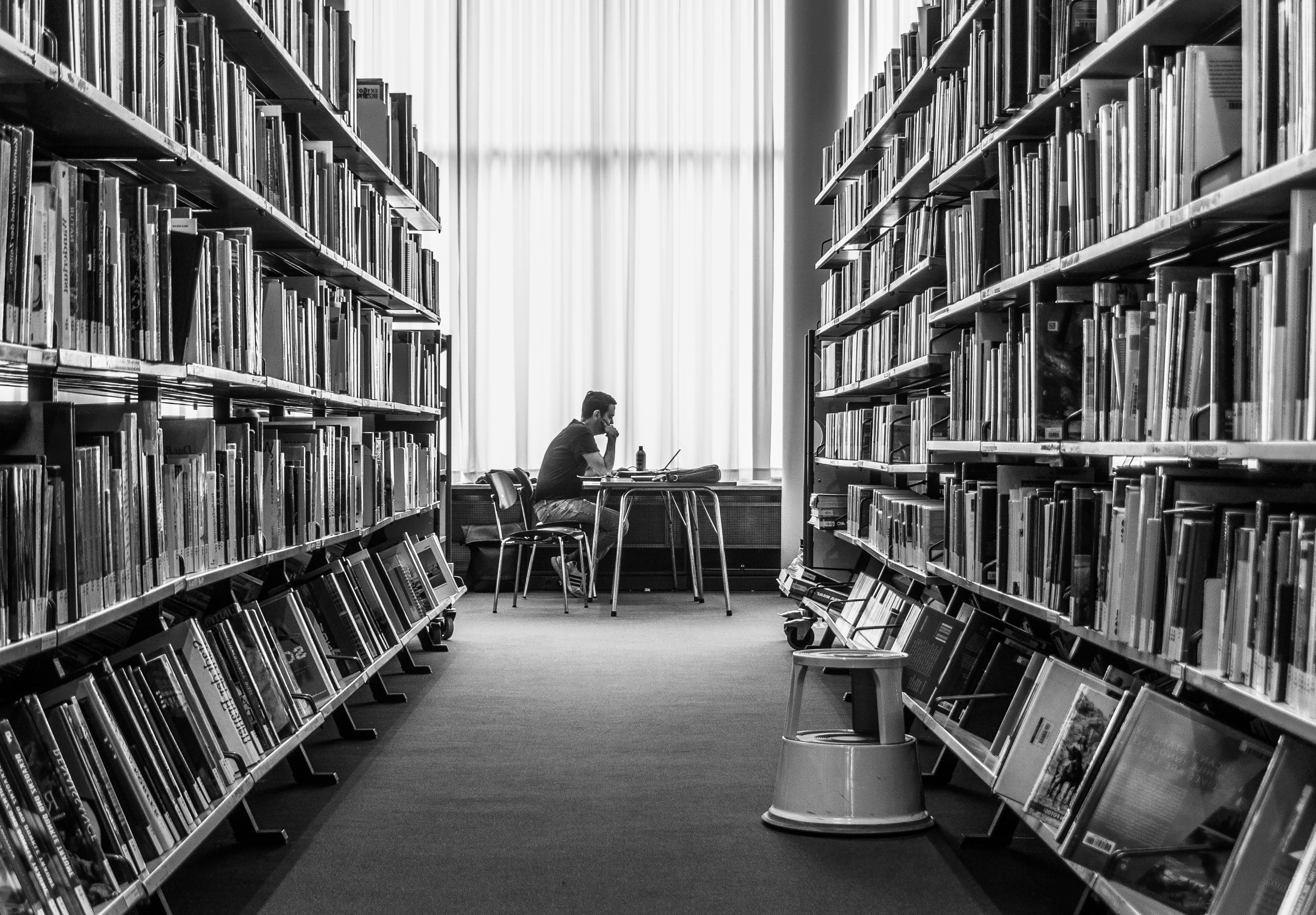 Man sitting beside a window in a library, seen through the space between two shelves lined with books